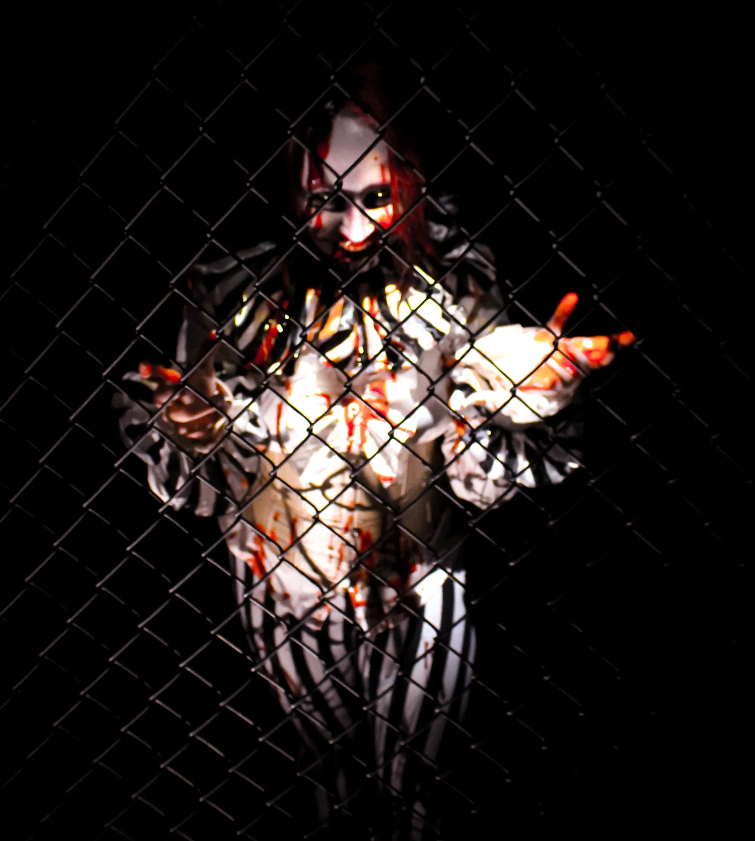 A person dressed as a creepy clown with a painted face, red hair, and a black and white costume, behind a wire mesh fence, pointing towards the camera with a dark background.