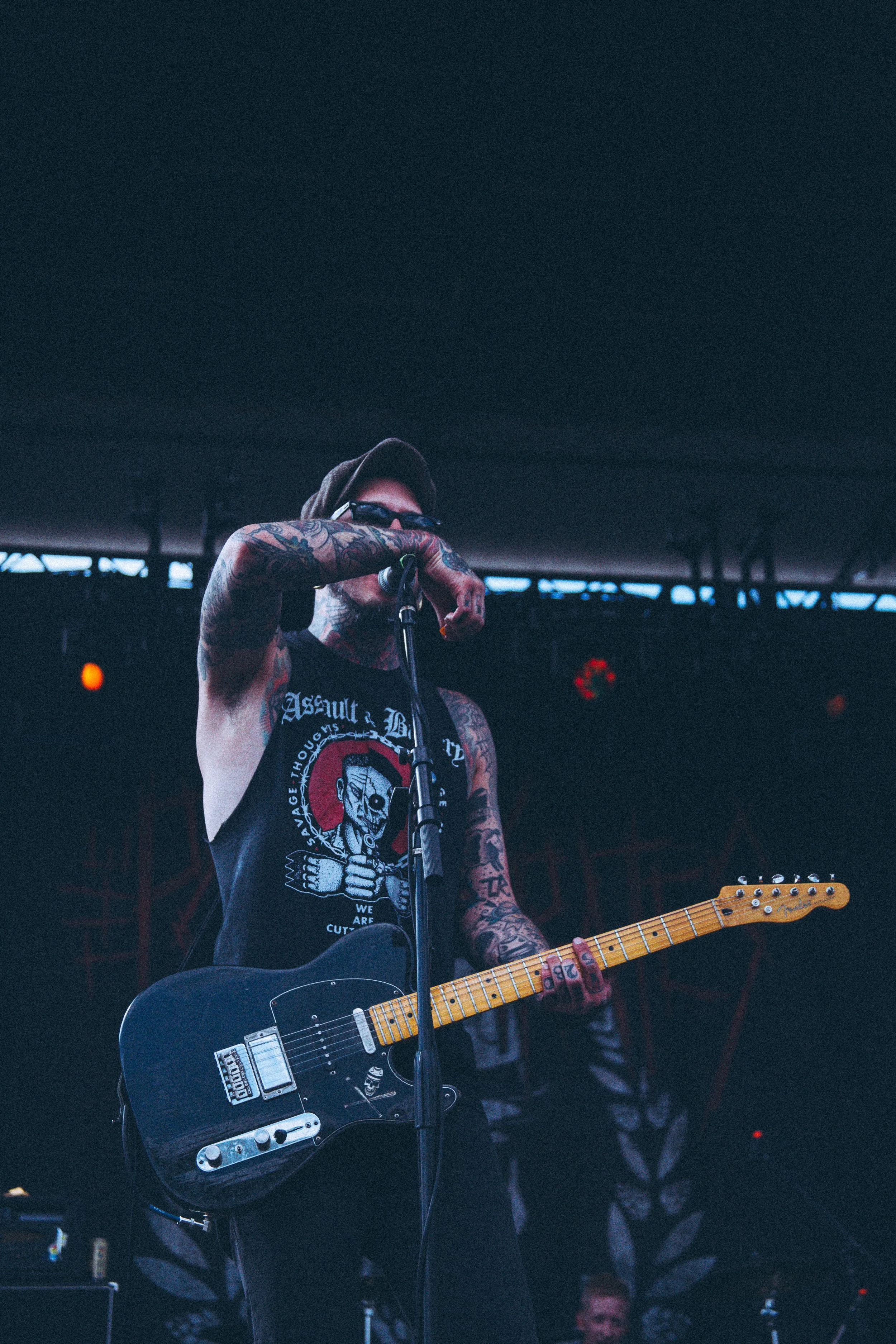 A tattooed male musician wearing sunglasses, a black sleeveless shirt, and a cap, singing into a microphone while playing an electric guitar on stage.