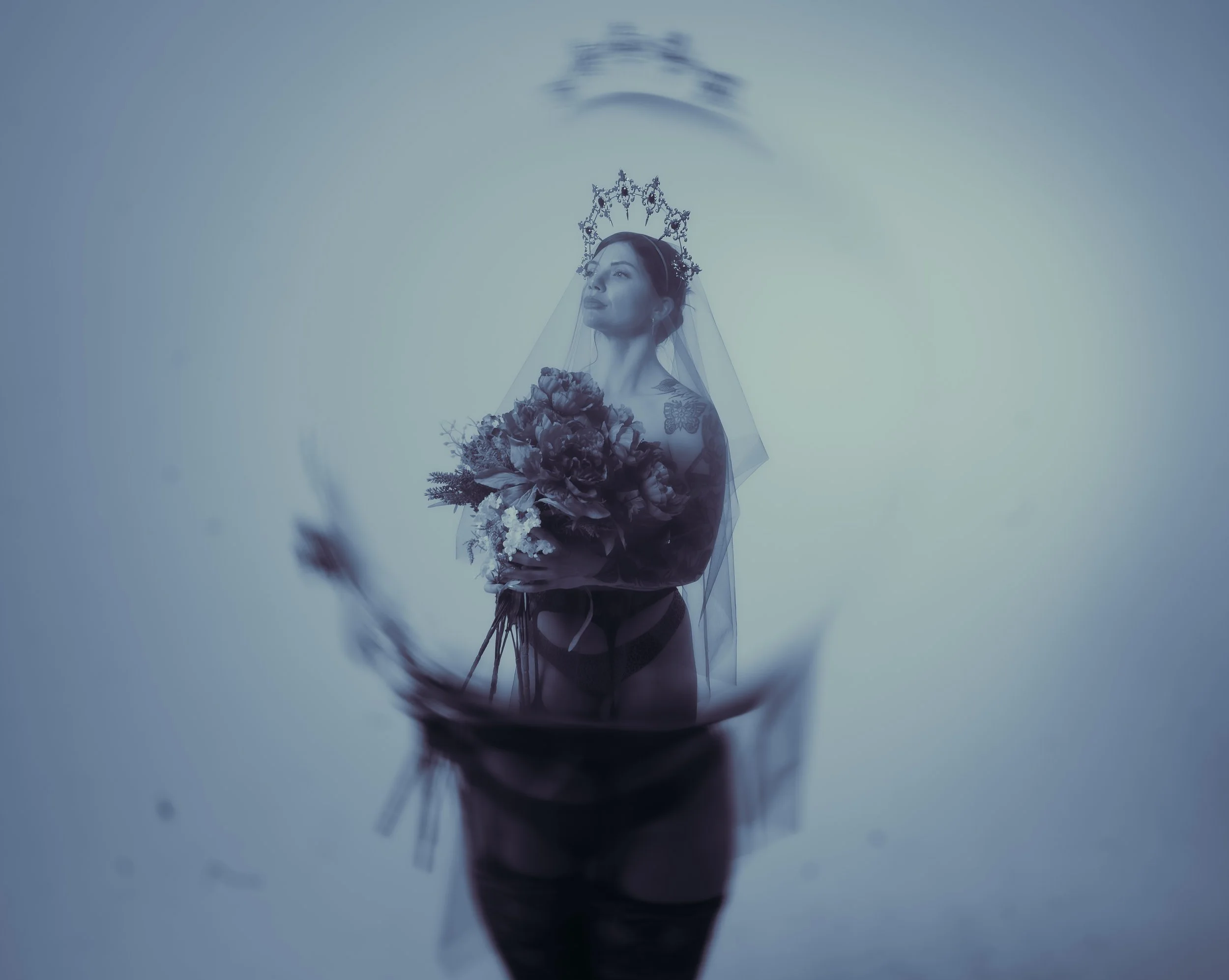 A woman in a bridal crown and veil holding a bouquet of flowers, reflected in water, with a blurred butterfly in the foreground.