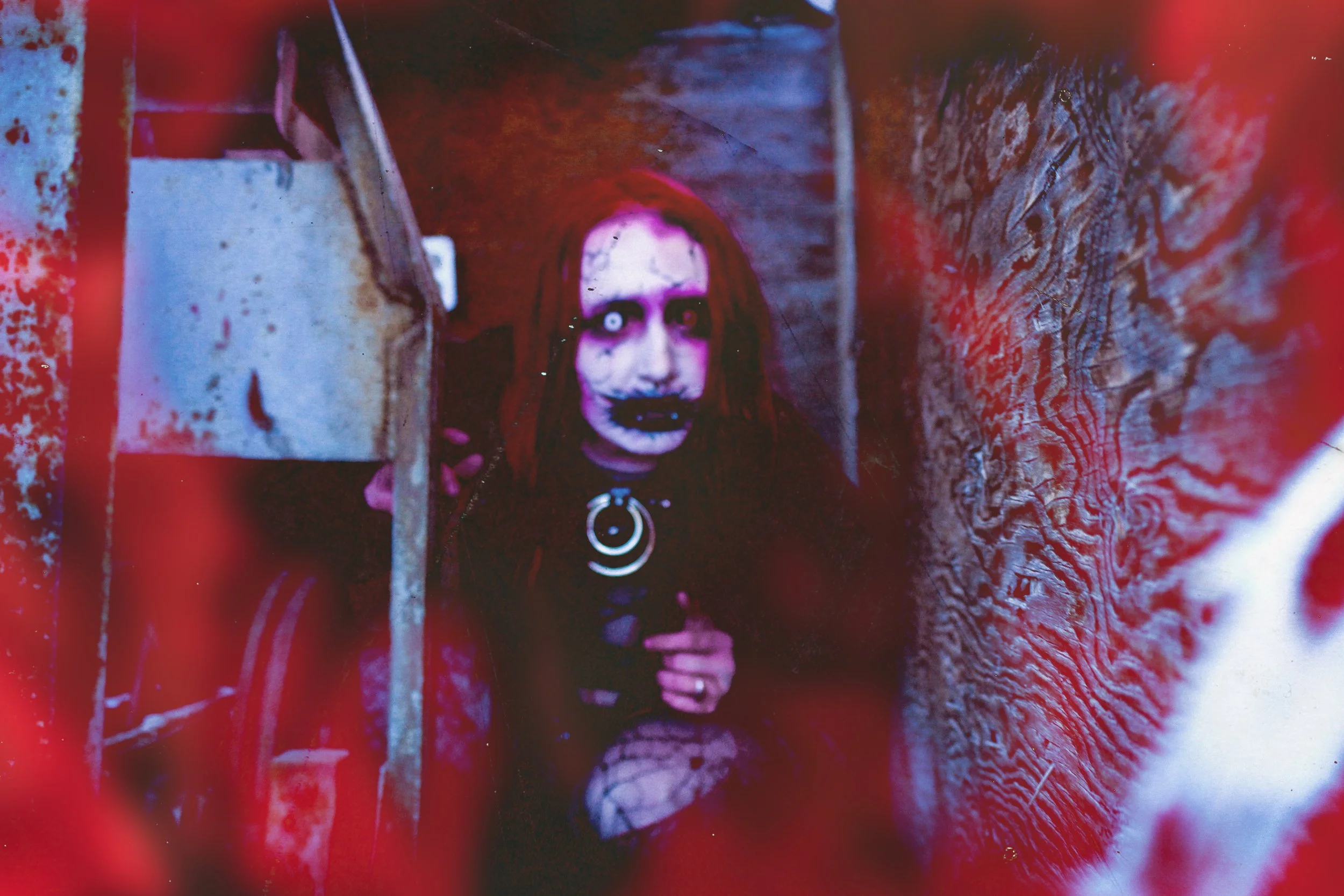 Person dressed in dark, goth-style clothing with corpse paint makeup, holding a flashlight, in a narrow, dark wooden corridor with red lighting.
