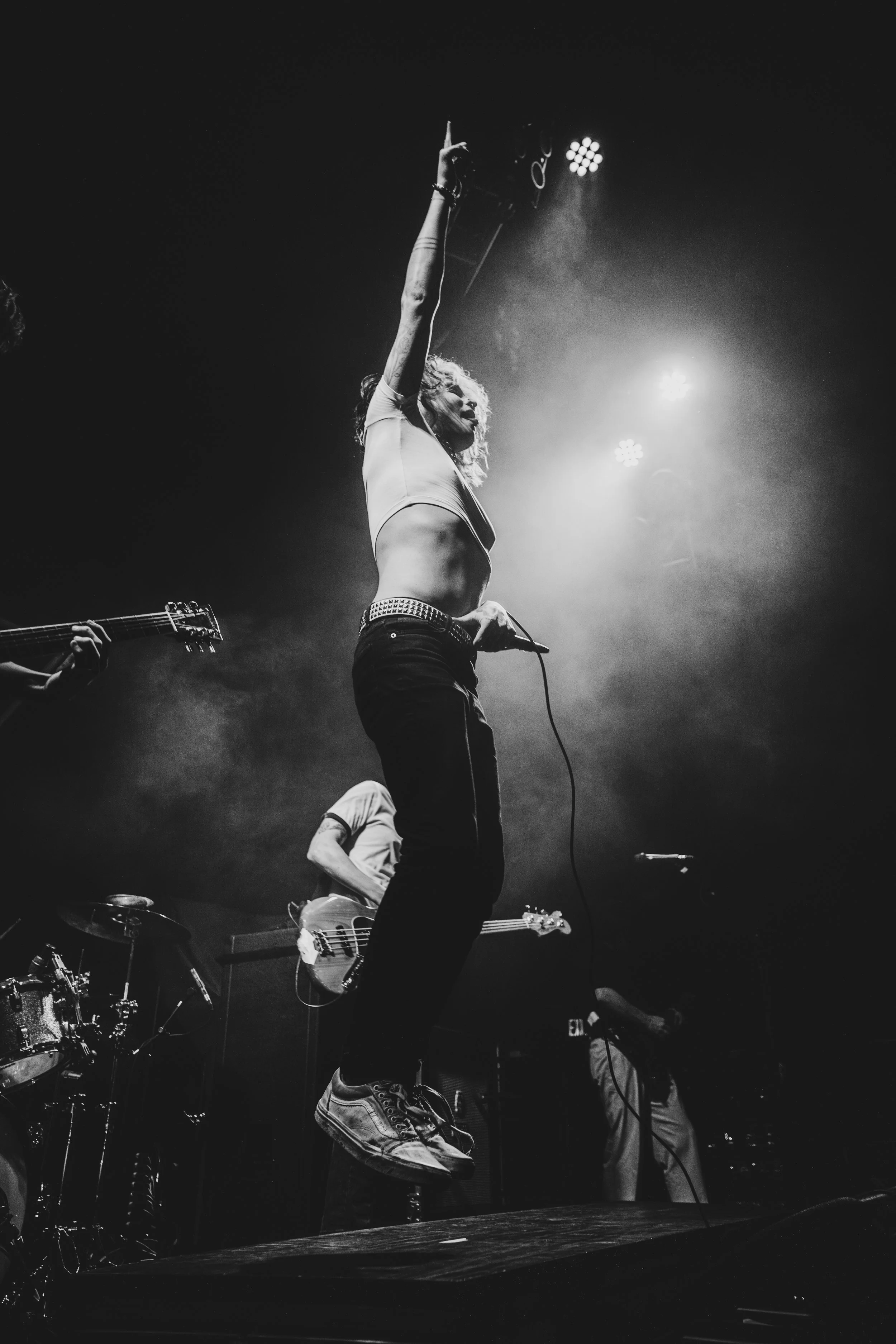 A singer with curly hair, wearing a crop top and jeans, jumps in the air while performing on stage, with band members playing instruments in the background, under stage lights.
