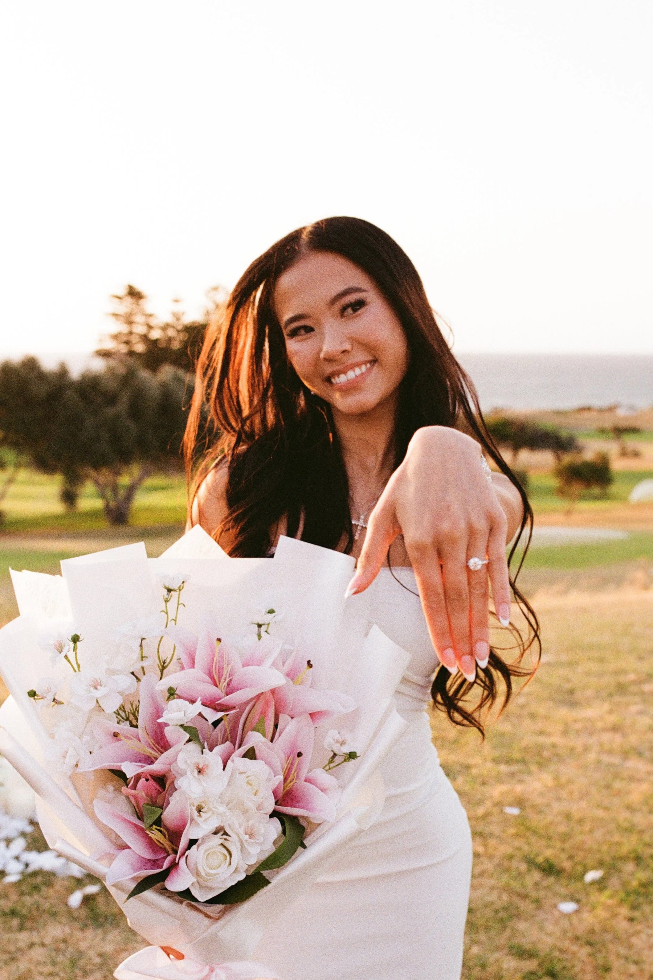 A woman in a white dress showing off an engagement ring on her finger while holding a bouquet of pink lilies and white flowers outdoors during sunset.