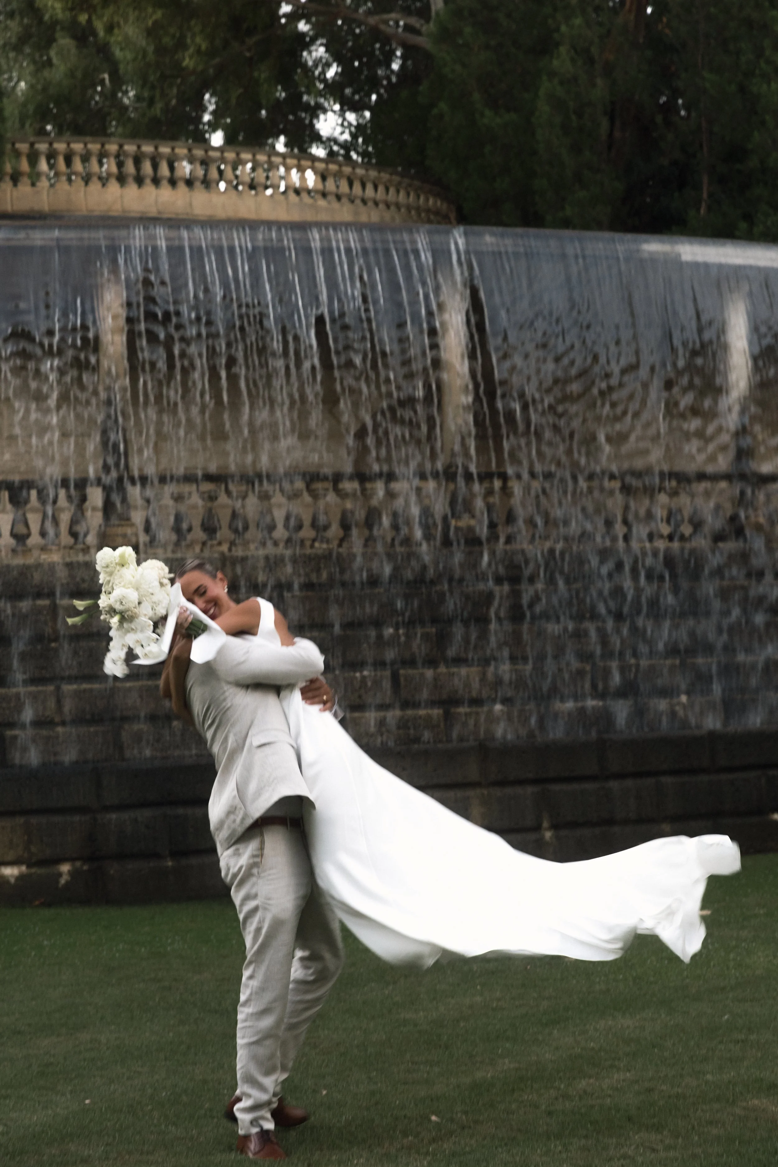 A wedding couple embracing near a fountain, with the bride holding a bouquet of white flowers and the groom lifting her, both smiling.