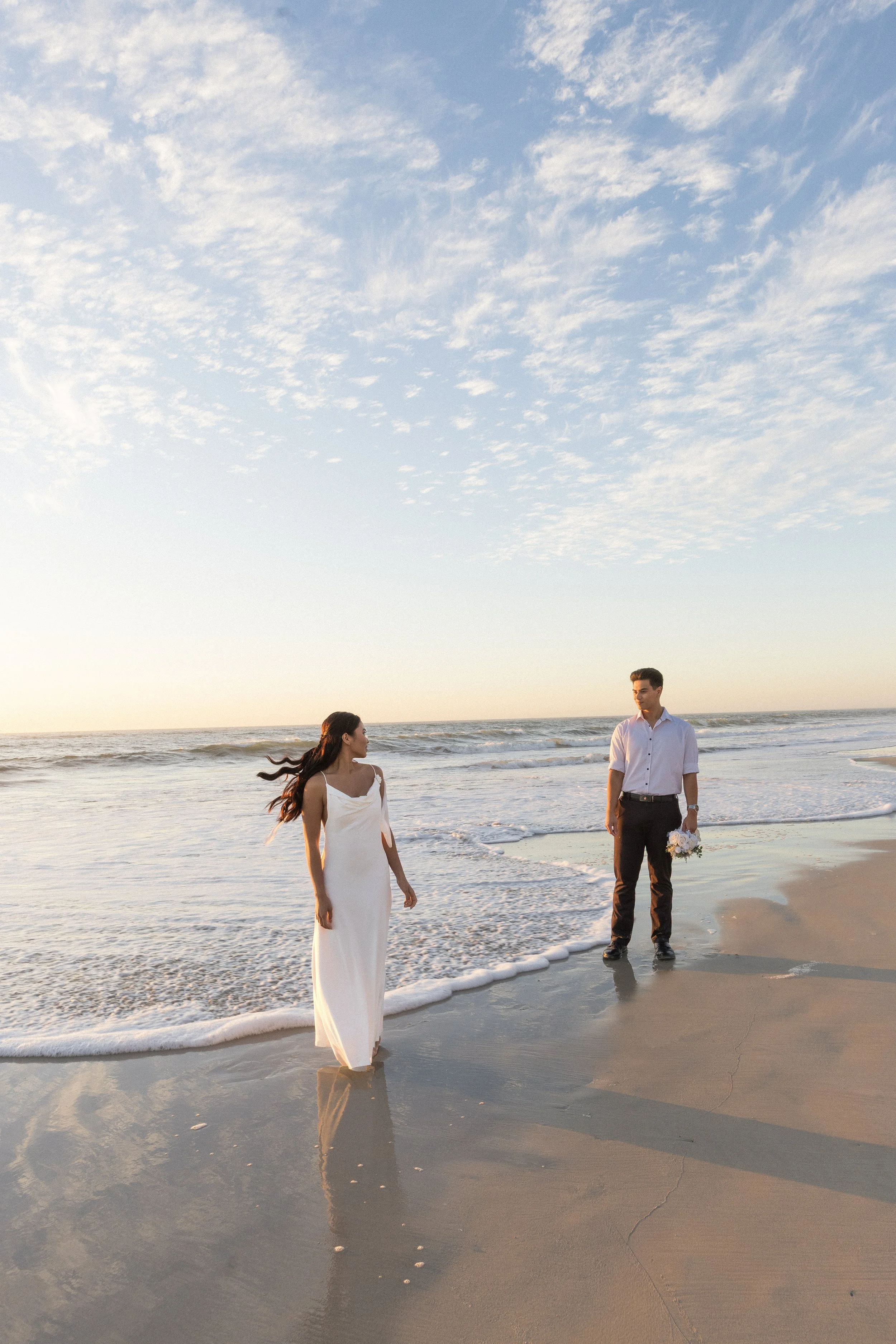 A couple dressed in wedding attire standing at the edge of the ocean on a sandy beach during sunset.