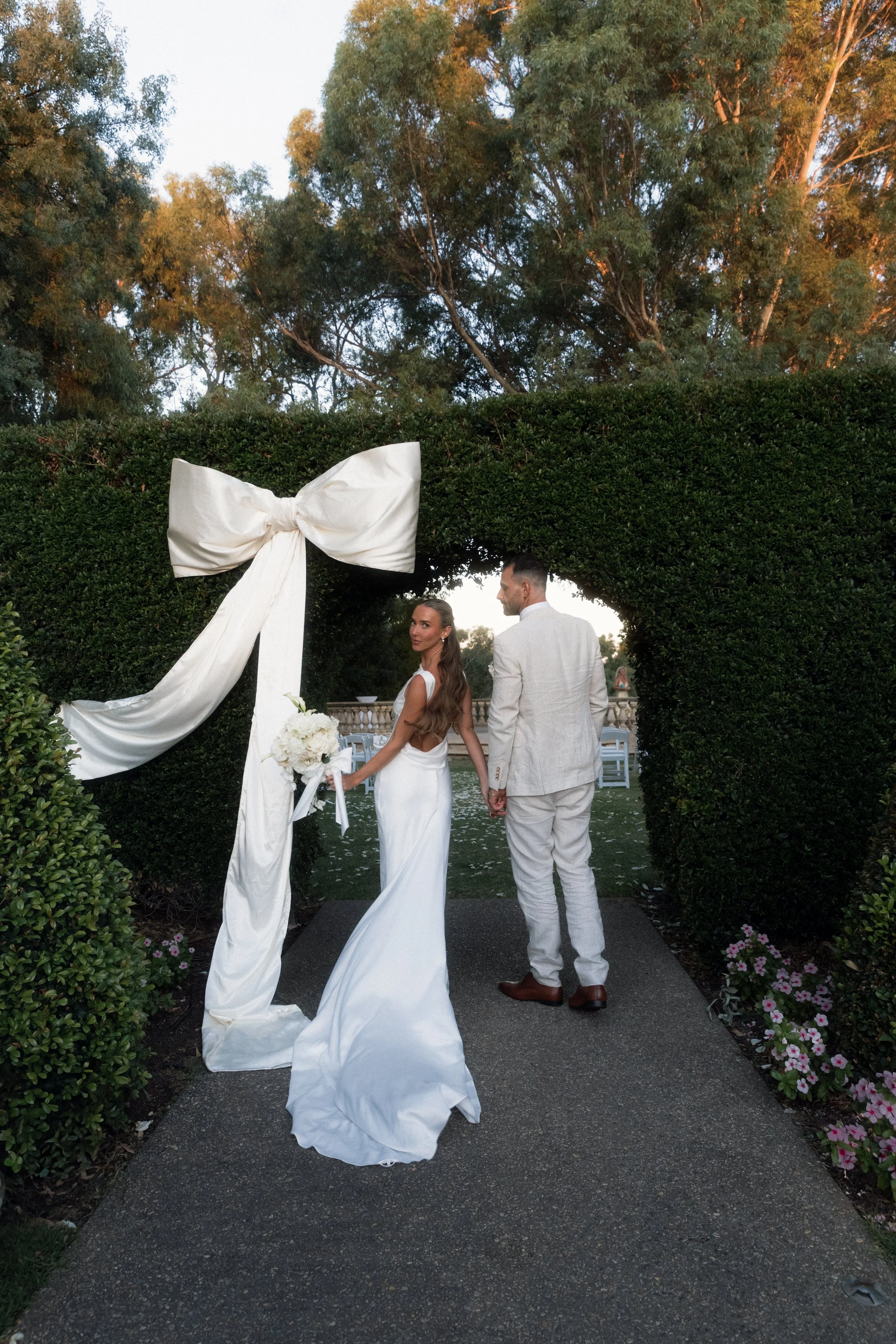 A bride and groom holding hands and walking through an archway decorated with a large white bow during their outdoor wedding ceremony.