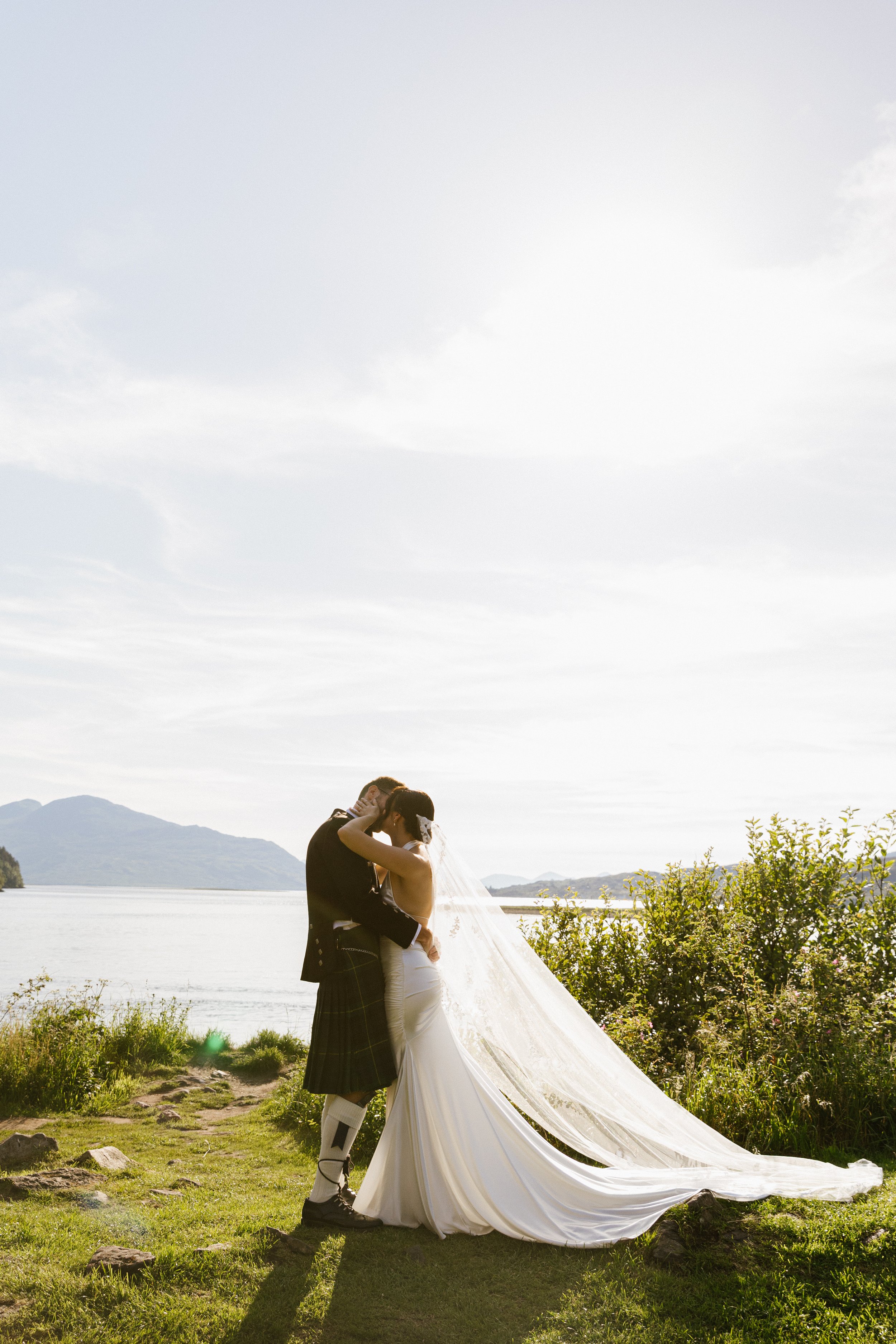 A bride and groom embracing outdoors on a grassy area near a body of water with the Scotland Highlands mountains in the background