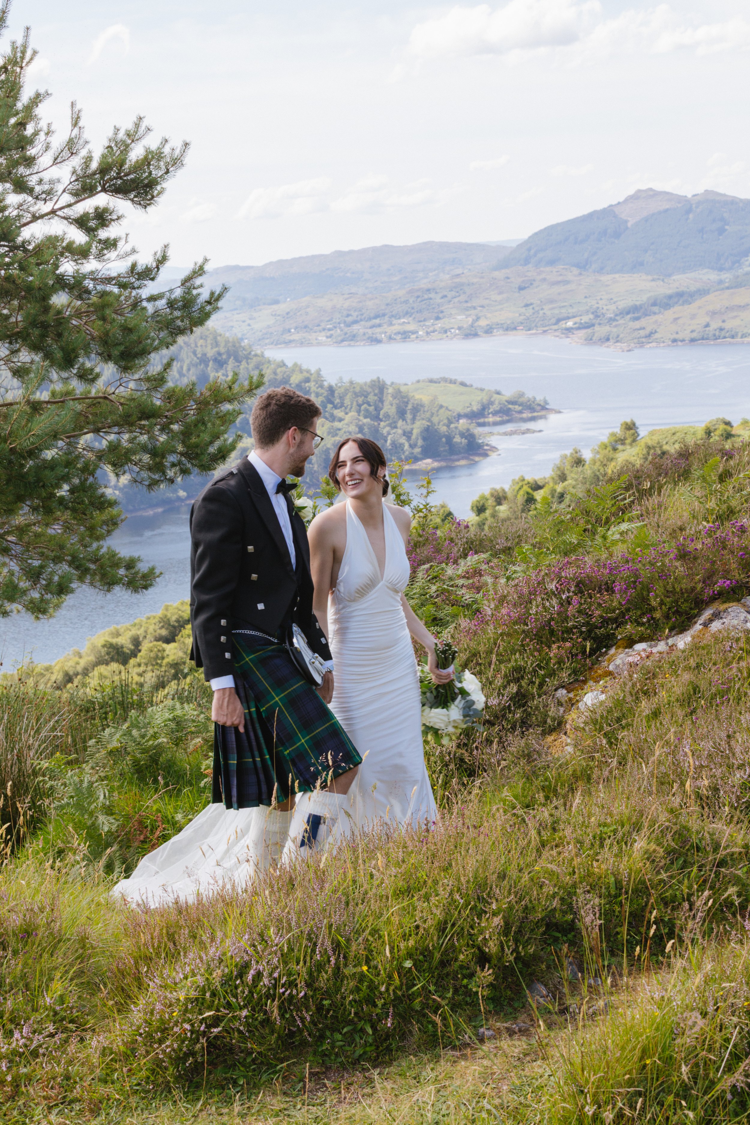 A newlywed couple walking outdoors on a grassy hillside with purple and green foliage, with the Scotland Highlands in the background. The bride is wearing a wedding dress and the groom is dressed in traditional Scottish attire, including a kilt.