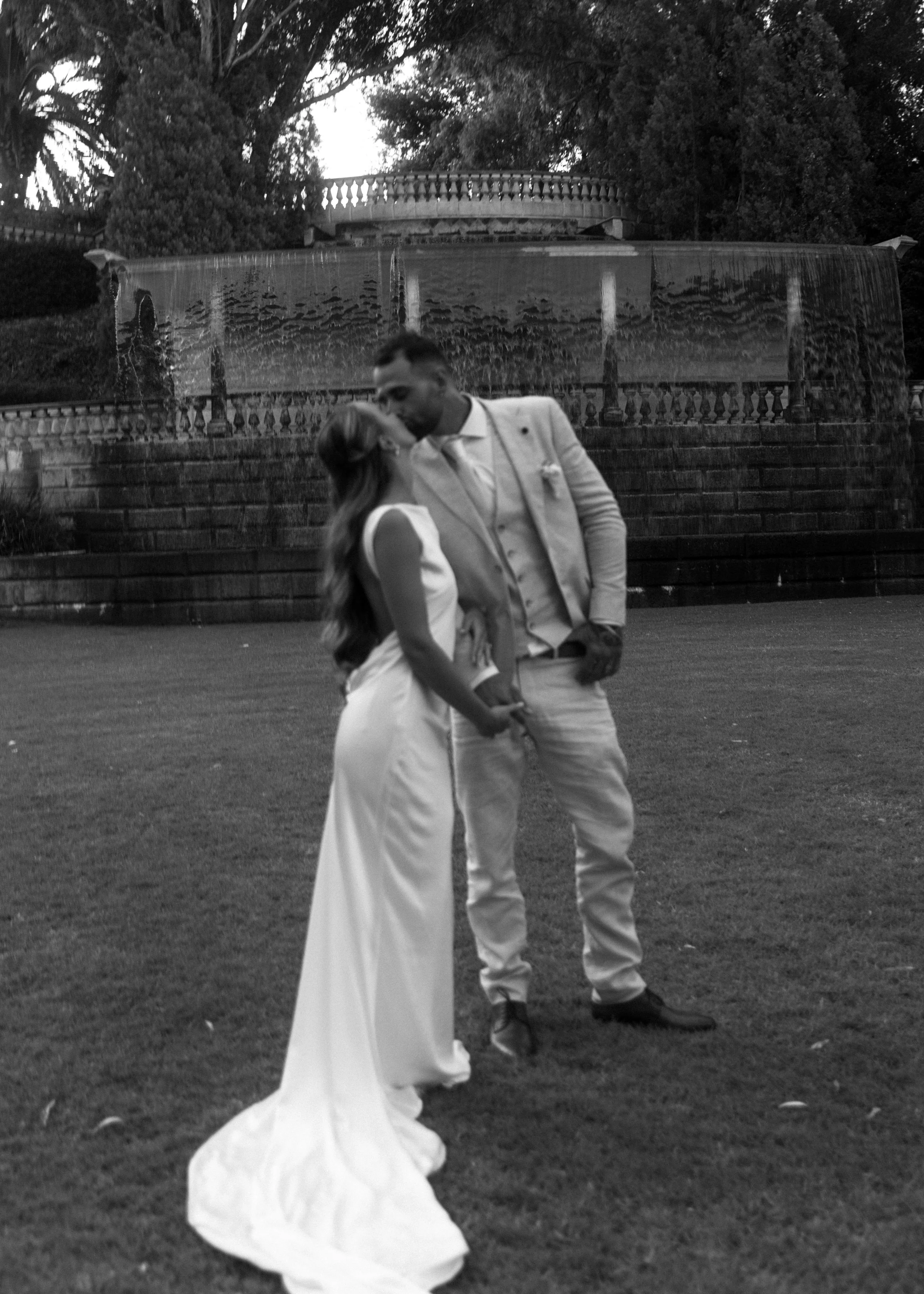 Black and white photo of a bride and groom sharing a kiss outdoors in front of a fountain, with trees and european railing in the background.