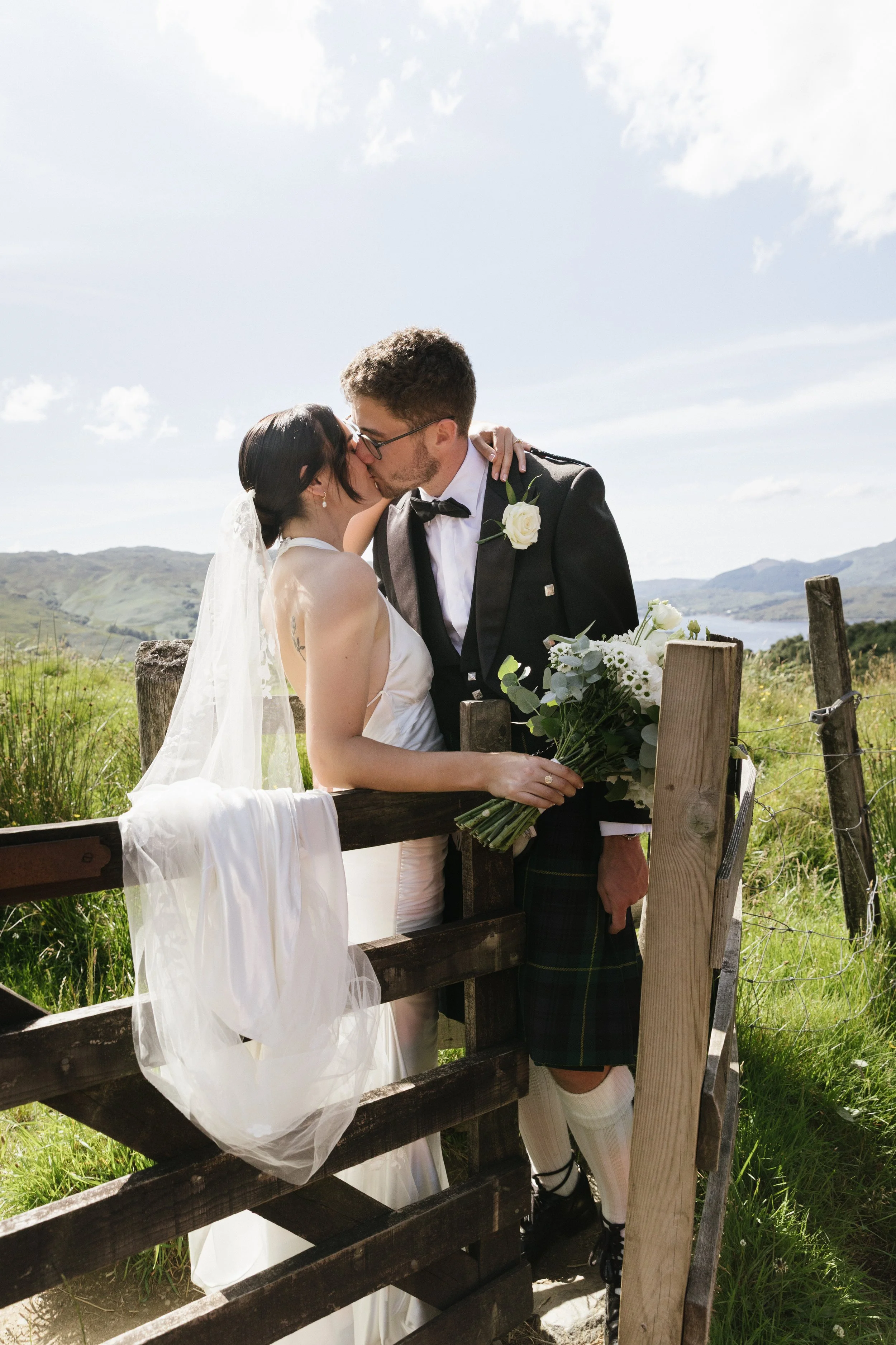 A bride and groom kissing outdoors on a sunny day, holding a bouquet of white flowers, standing by a wooden fence in the Scotland Highlands.