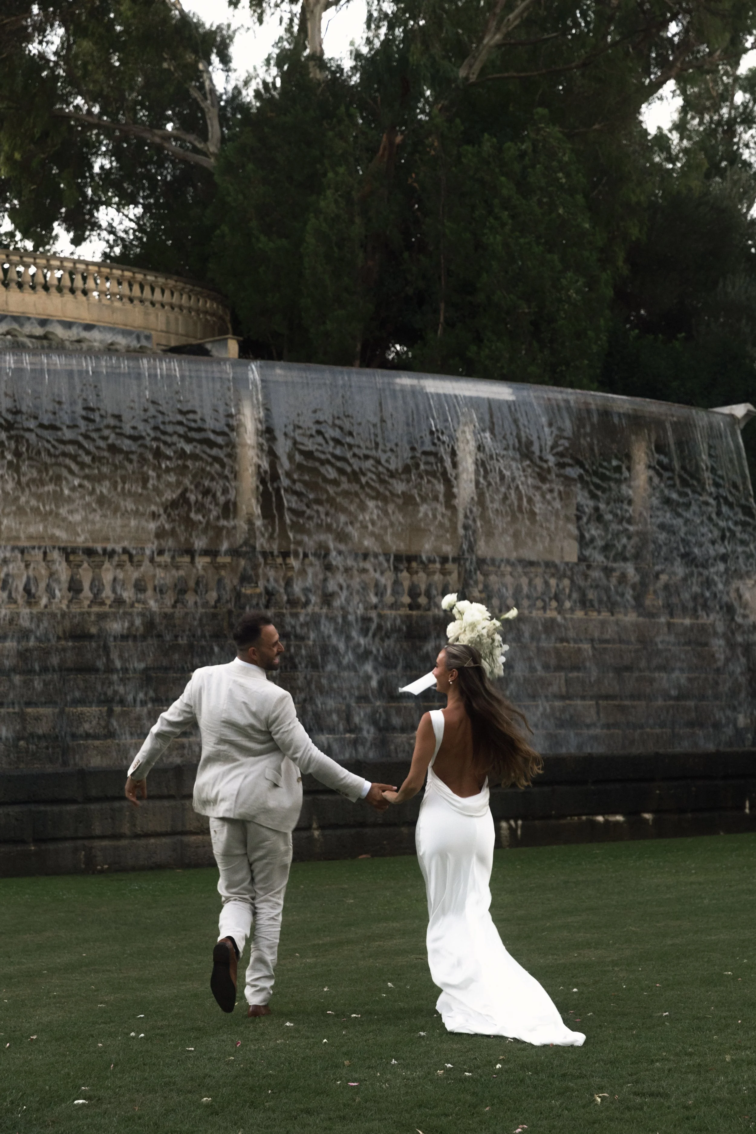 A bride and groom holding hands, running on a grassy area in front of a large stone waterfall fountain, with trees in the background, during a wedding or romantic celebration.