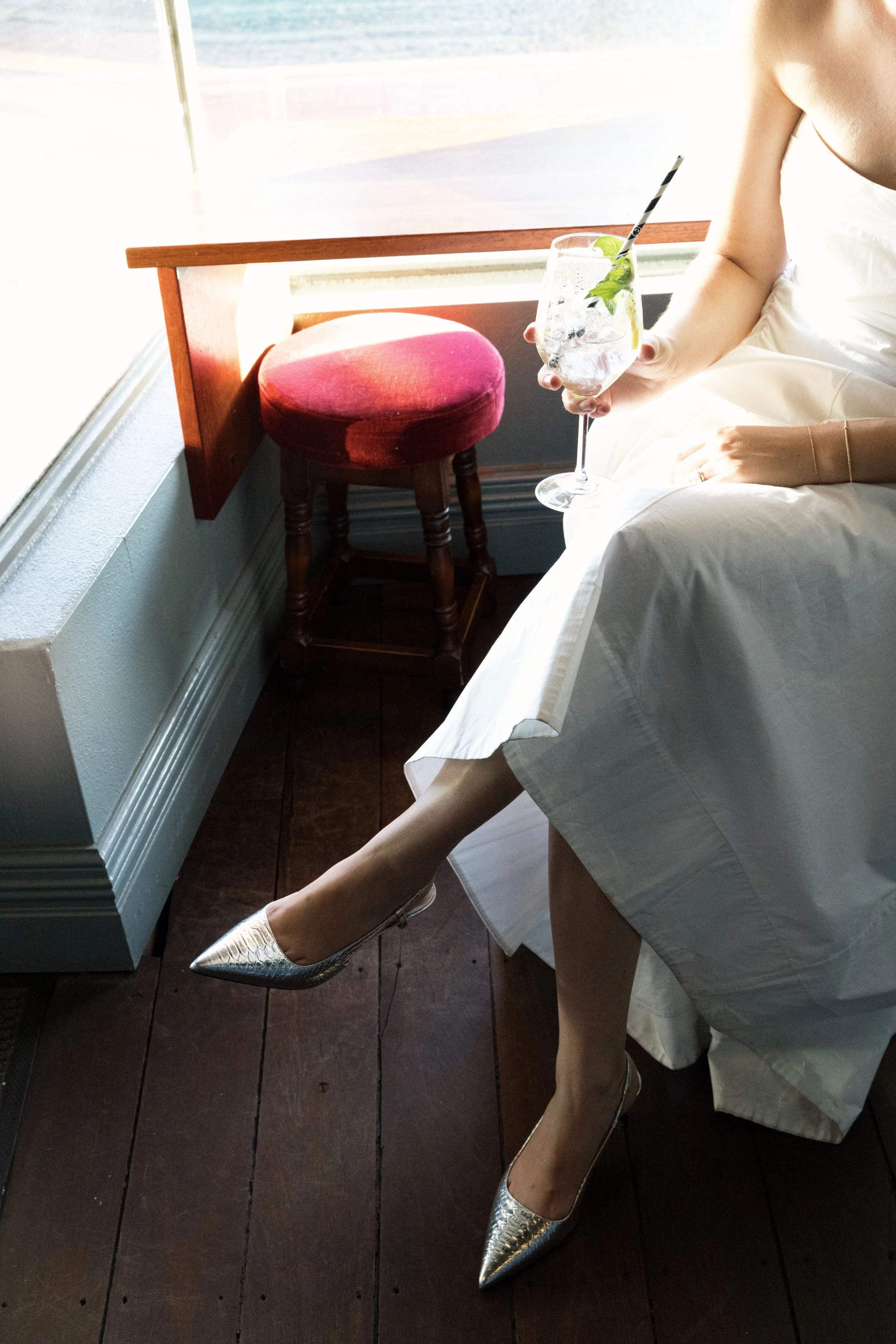 A woman holding a cocktail: Hugo Spritz, with a straw and mint leaves, sitting on a bed by a window with sunlight, wearing a white dress and silver high heels.