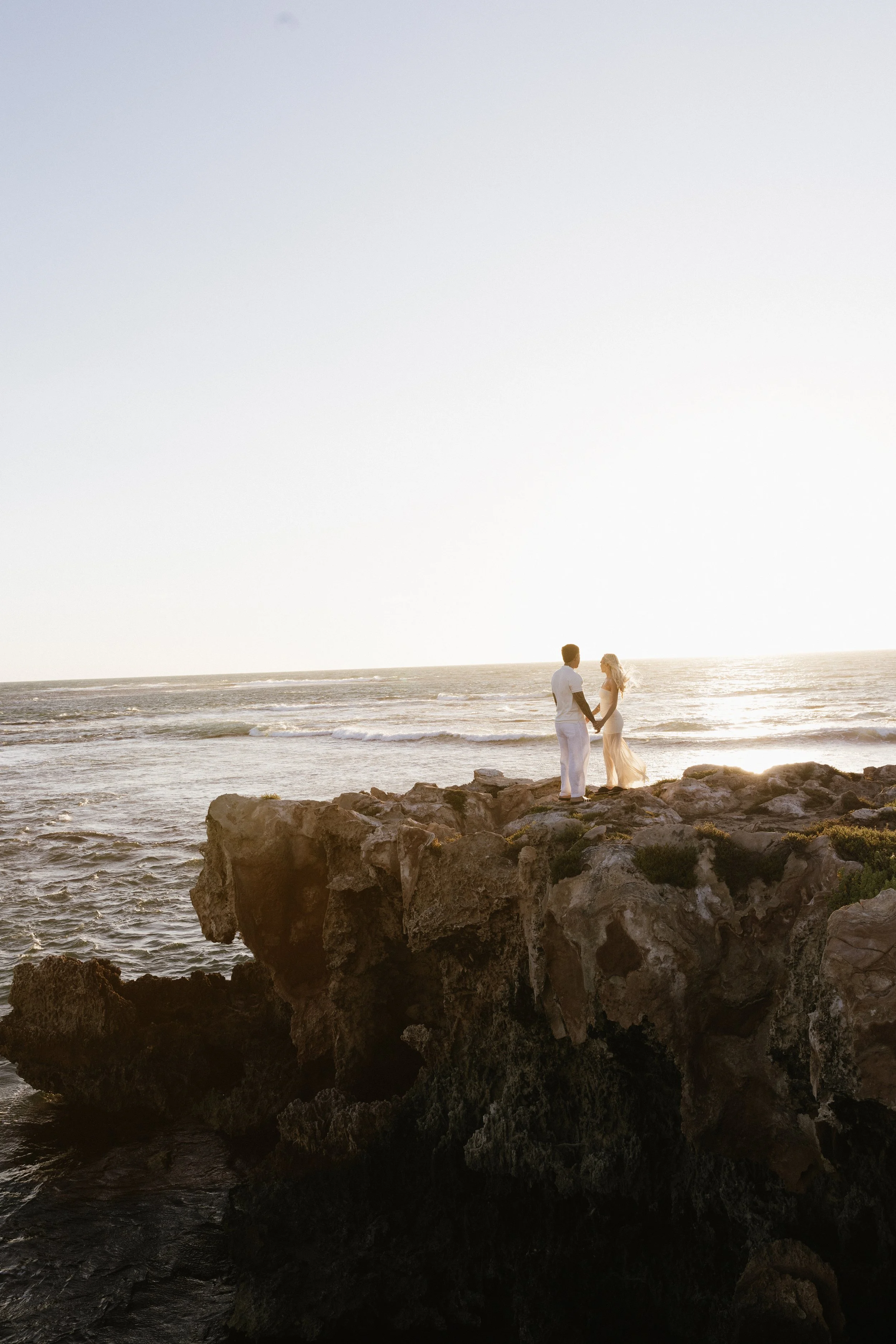 A couple in wedding attire standing on a rocky cliff by the ocean during sunset, holding hands and facing each other.