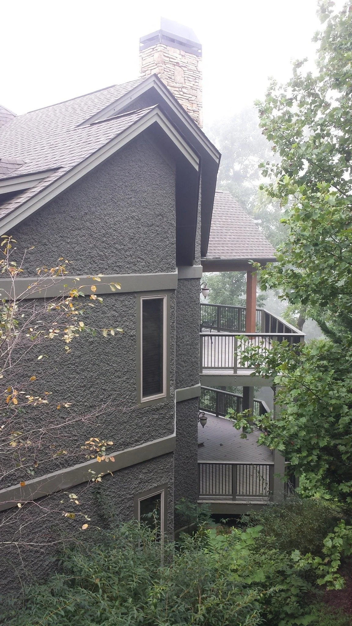 A multi-story house with textured gray exterior walls, surrounded by trees and partially obscured by fog. The house has a gabled roof, a brick chimney, and outdoor balconies with black railings.