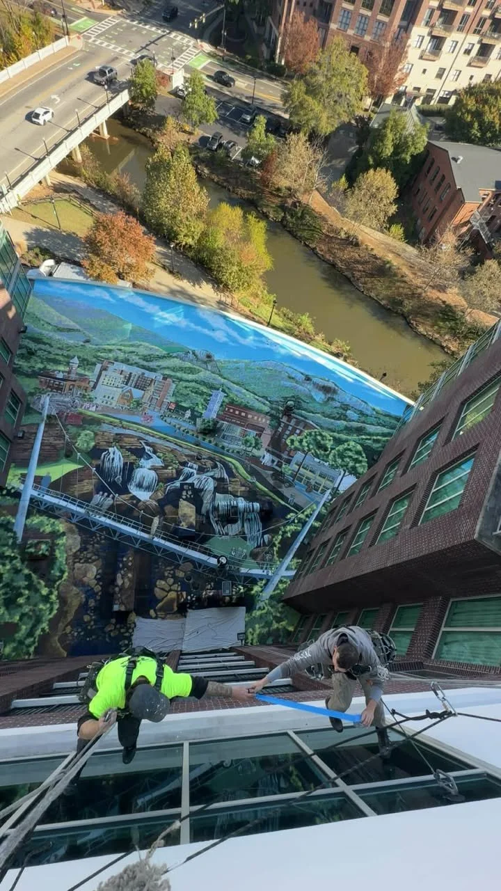 Two workers in safety gear climbing a tall building's exterior, with a mural of a cityscape and river below them.