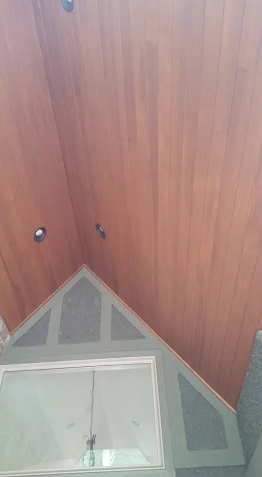 A corner of a room showing a wood-paneled wall with two circular electrical outlets or switches and a ceiling with a hanging light fixture, reflected in a large mirror on the lower wall.