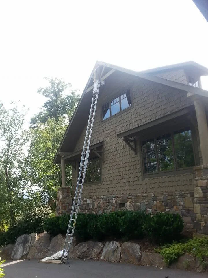 A ladder leaning against the corner of a two-story house with stone and siding exterior, under a bright sky.