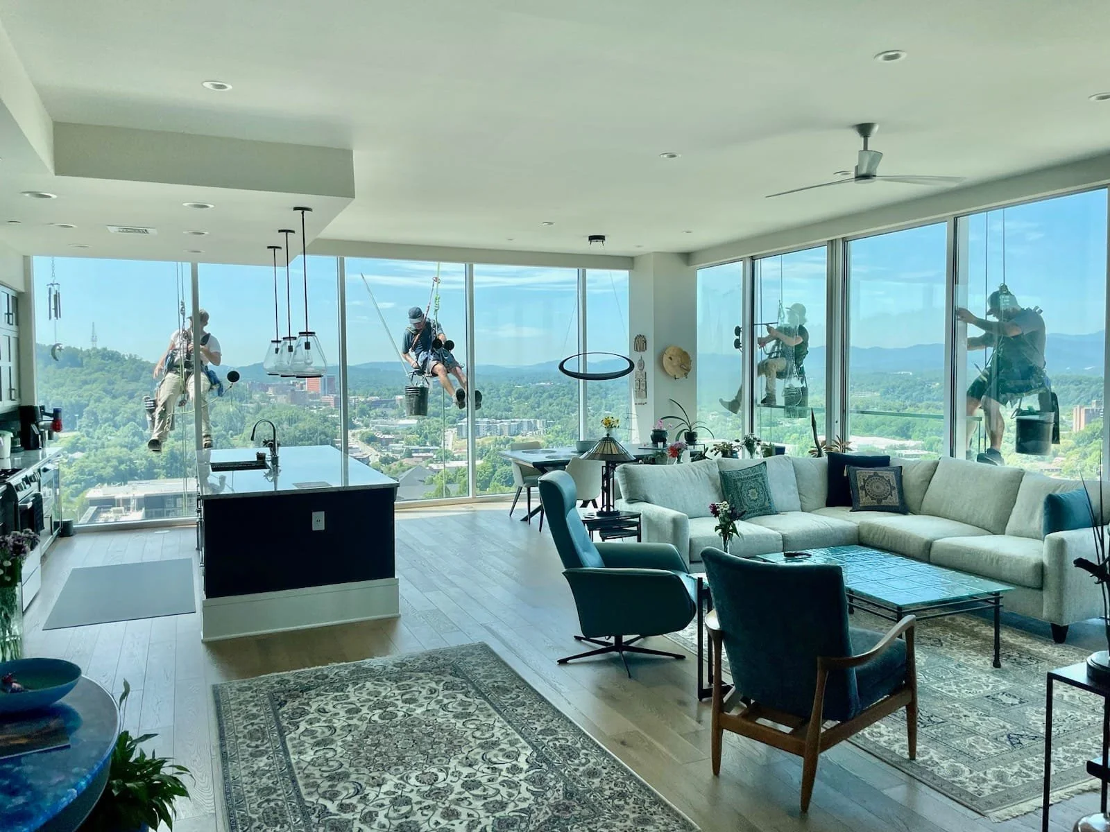Living room with large windows showing city view and five workers cleaning exterior windows with harnesses.