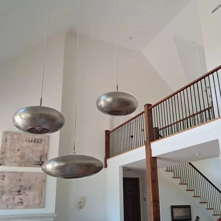 Three large metal pendant lights hanging from the ceiling in a modern home with high ceilings, a wooden railing on the upper level, and a staircase leading downstairs.