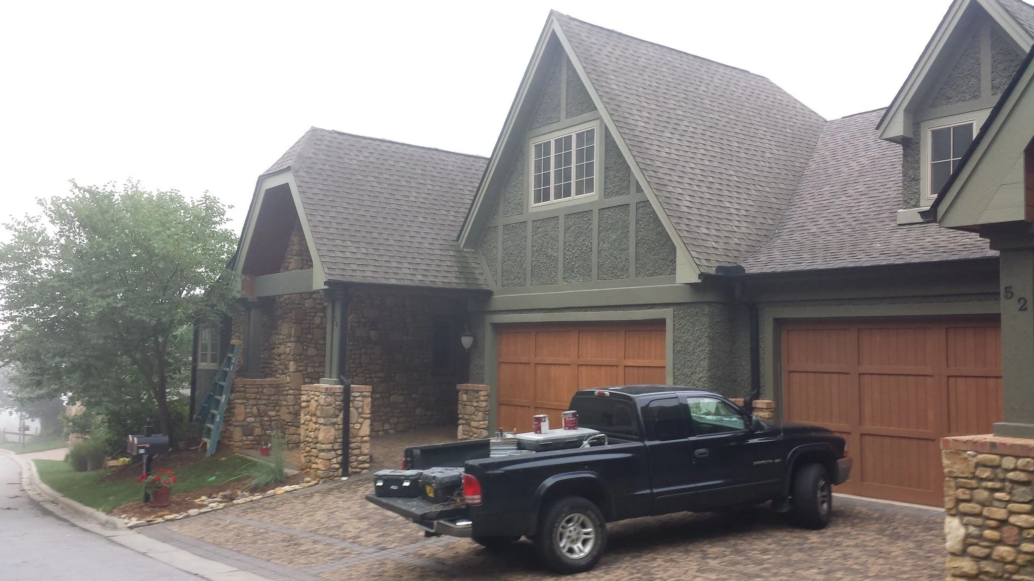 Front view of a large house with a stone and stucco exterior, a wooden garage door, and a black pickup truck parked in the driveway. Tree and foggy weather in the background.