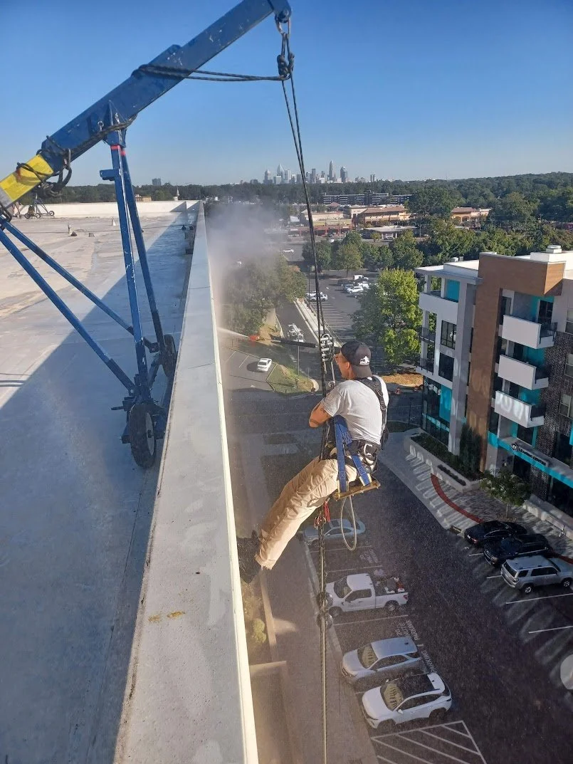 A worker sitting on a harness and safety rope on a rooftop edge, using a pressure washer to clean the concrete of a high-rise building, with a city skyline in the background.
