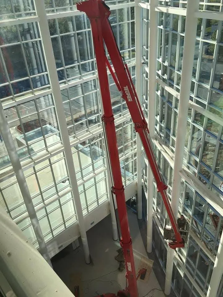 A red telescopic boom lift inside a multi-story glass building with white framework, extending toward the upper floors to perform maintenance or cleaning.