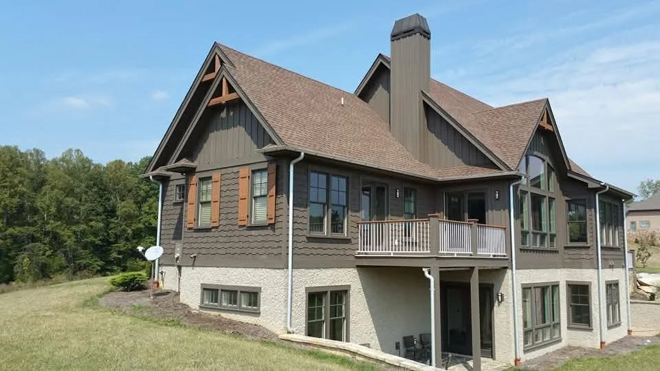 A large two-story house with dark gray siding, brown roof, multiple windows, and a small balcony on the second floor, set against a blue sky and green trees.
