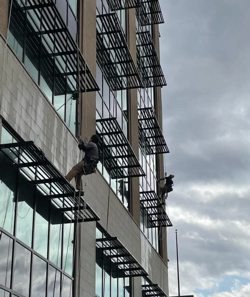 Two window cleaners with water-fed poles, WFP,  washing windows on the exterior of a tall modern building with multiple glass windows and metal awnings.