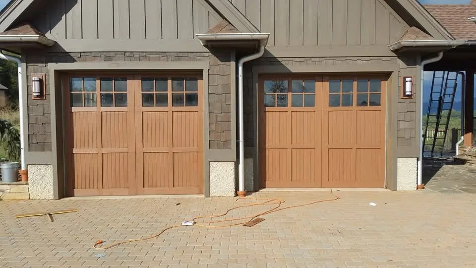 Two new garage doors made of wood with a grid of windows at the top, set in a house with gray siding and stone accents.