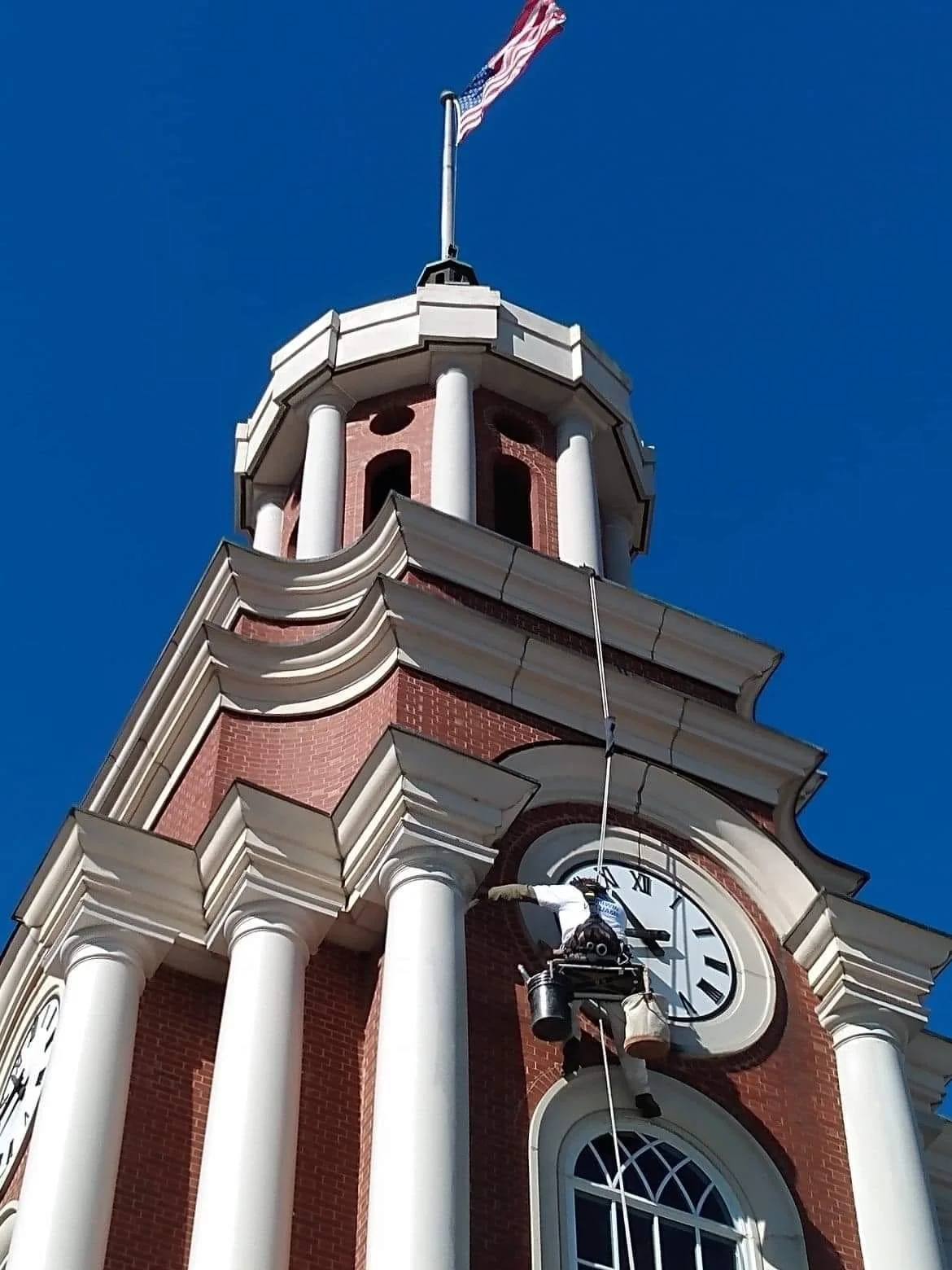 A person cleaning the clock on a tall red brick building with white columns, an American flag on top, and a clear blue sky in the background.