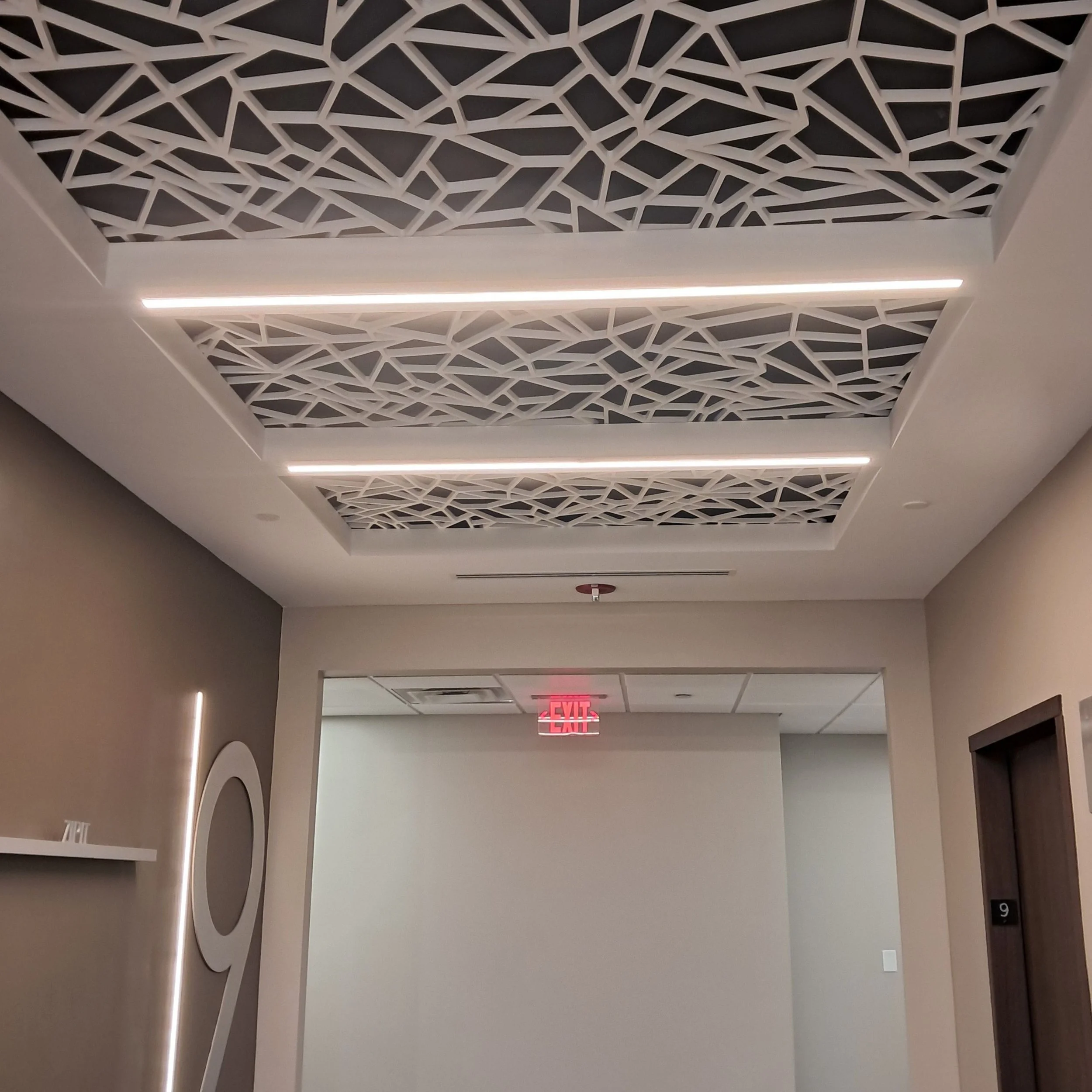 Modern interior ceiling with geometric patterned panels and integrated linear lighting, with an exit sign visible in the background.