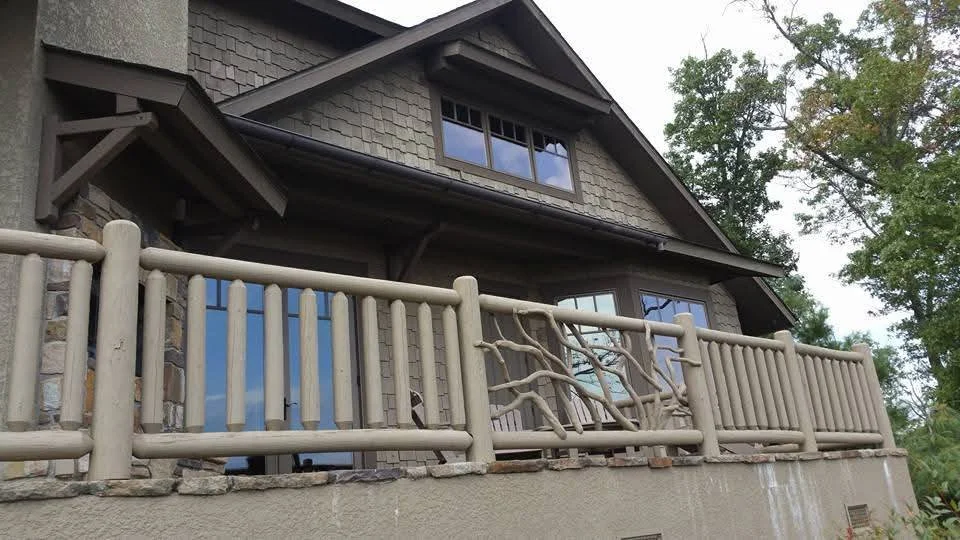 A house with a stone exterior and a wooden balcony railing, surrounded by tall trees.
