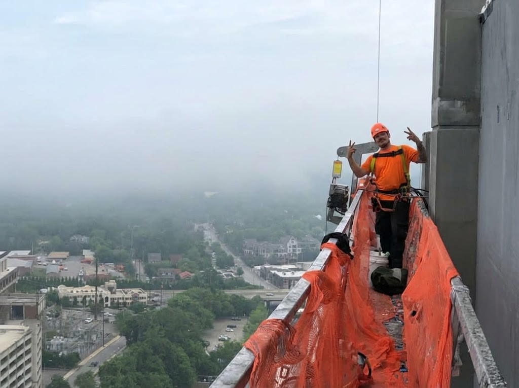 A construction worker standing on a narrow metal platform on the side of a tall building, wearing orange safety gear and helmet, making peace signs with both hands, with a misty cityscape in the background.