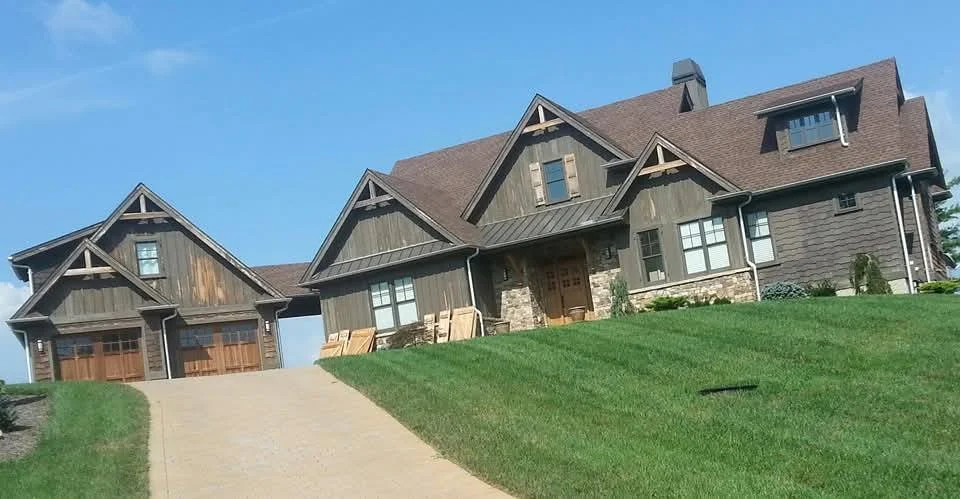 Large house with wood and stone exterior, multiple gabled roofs, attached garage, and well-maintained lawn with a concrete driveway.