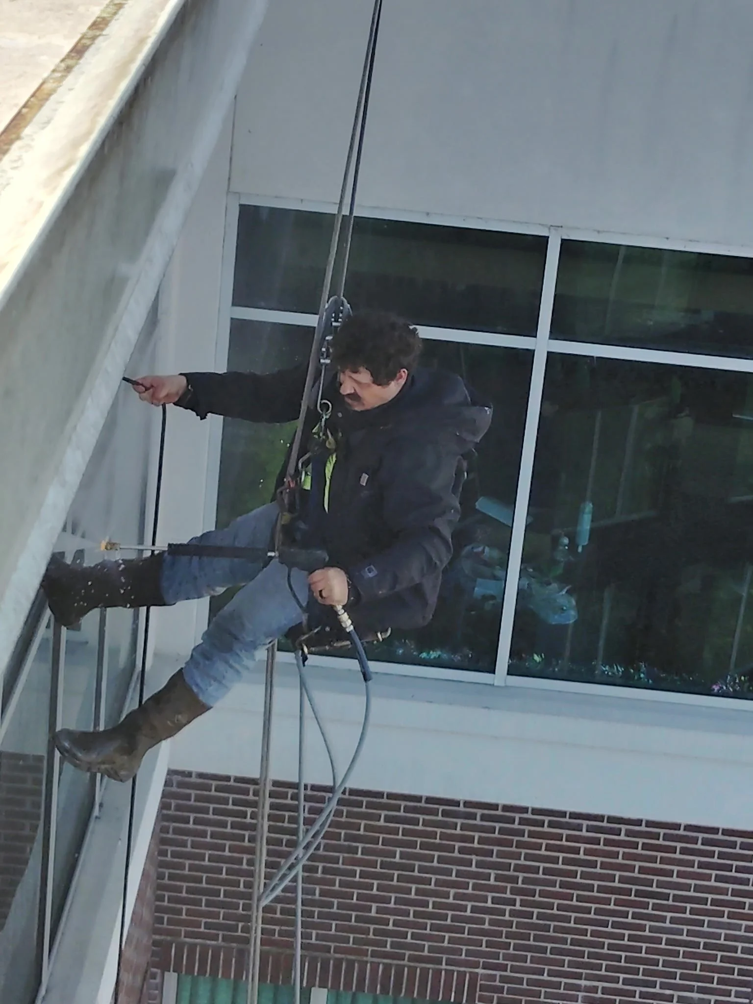A man is rappelling down the side of a building pressure washing using safety harnesses and ropes, wearing boots and outdoor clothing.