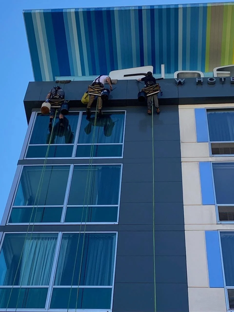 Three window washers hanging from ropes and cleaning the exterior of a tall building with large glass windows and a hotel sign at the top.