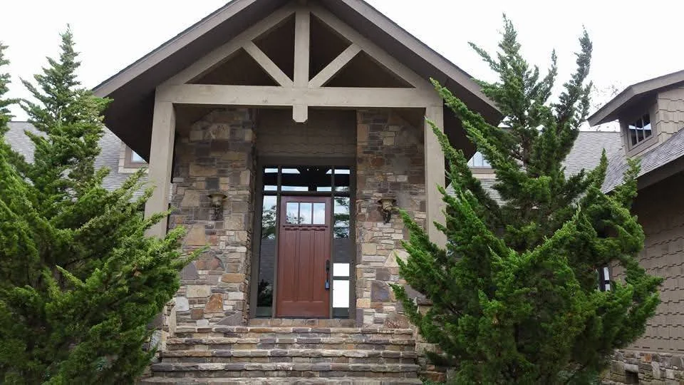 Front view of house with stone stairs, wooden front door, small square window, and surrounding green trees.