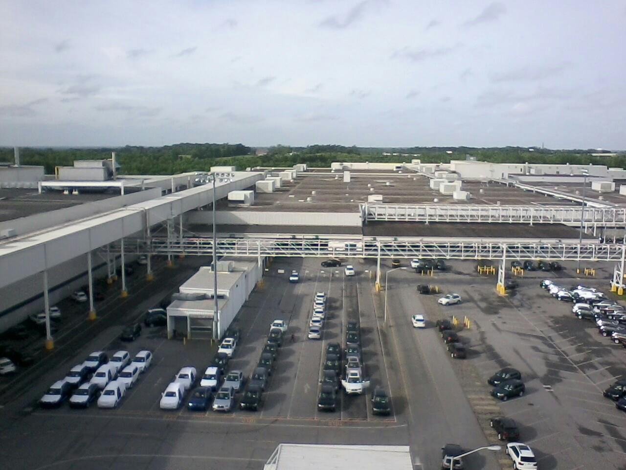 View of a large commercial building's rooftop and parking lot with cars parked in rows, overcast sky, green trees in the distance.