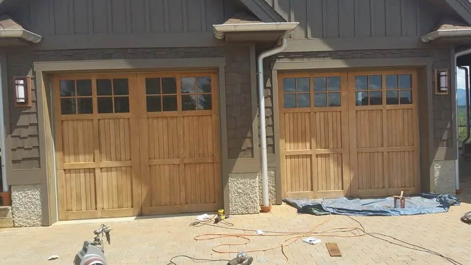 Two wooden garage doors with window panels at the top, installed on a house with grey siding and stone accents. Construction tools, extension cords, and tarps are on the ground in front of the doors.