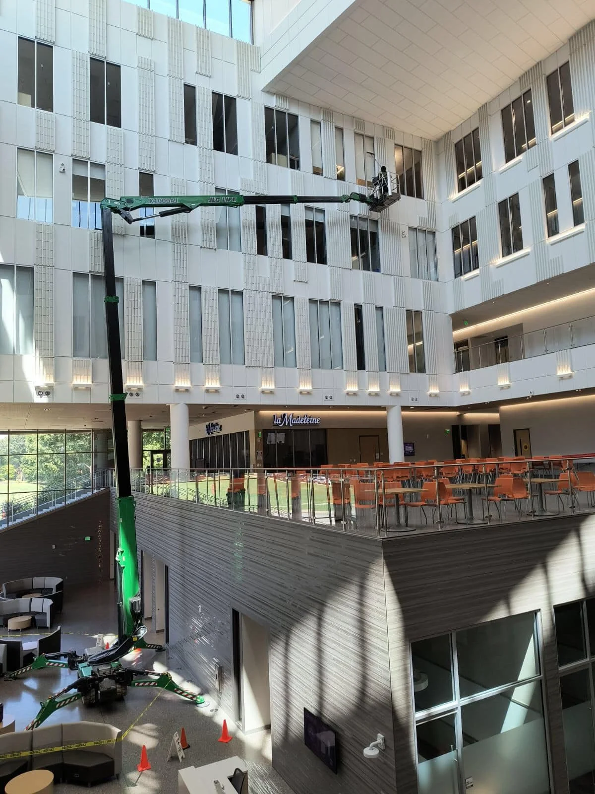 Interior of a modern multi-story building with a large indoor space. A green hydraulic lift is extended towards the upper floor, with a worker inside the lift working on the windows or ceiling. The upper floor has a lounge area with orange chairs and tables, with a sign that reads 'la Madeleine' in the background. The sunlight streams through large windows onto the lower level, where there are some seating arrangements and orange traffic cones.