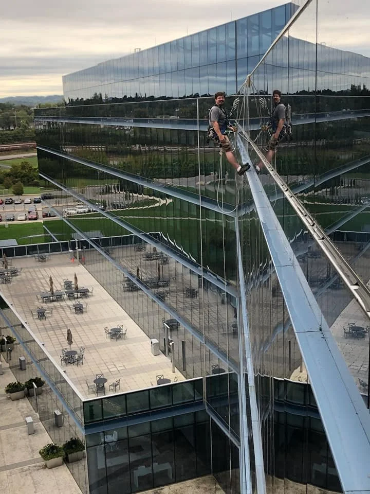 Two window washers with safety harnesses cleaning the windows of a skyscraper. They are sitting on a suspended platform high above the ground, reflected in the glass.