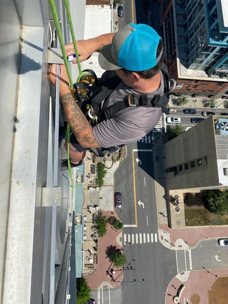 A worker securing a safety harness on a tall building's edge during construction, high above city streets with cars, trees, and sidewalks.