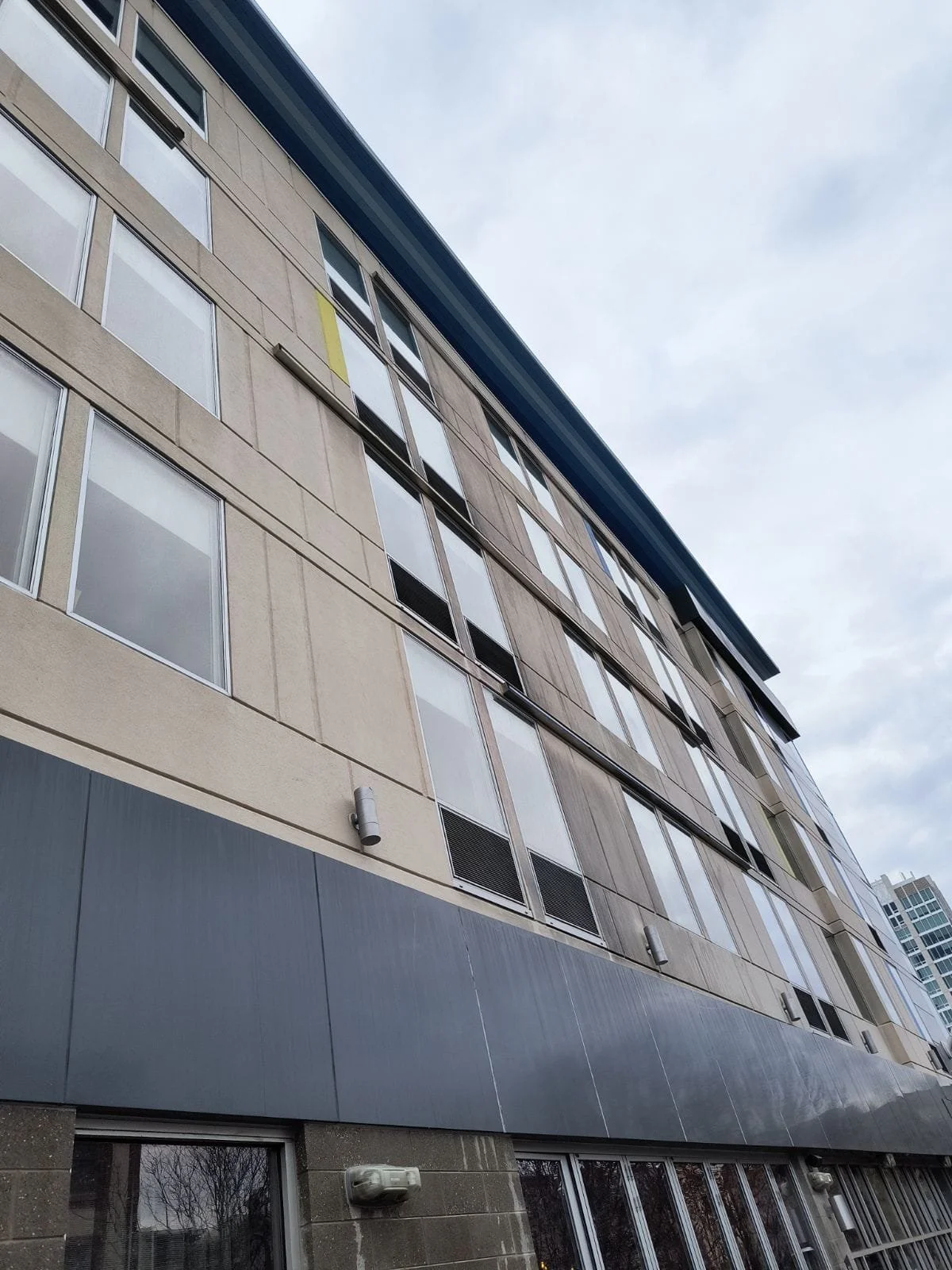 A multi-story modern building viewed from below, showing numerous windows and a mix of beige and gray exterior walls, with some small exterior lights visible.
