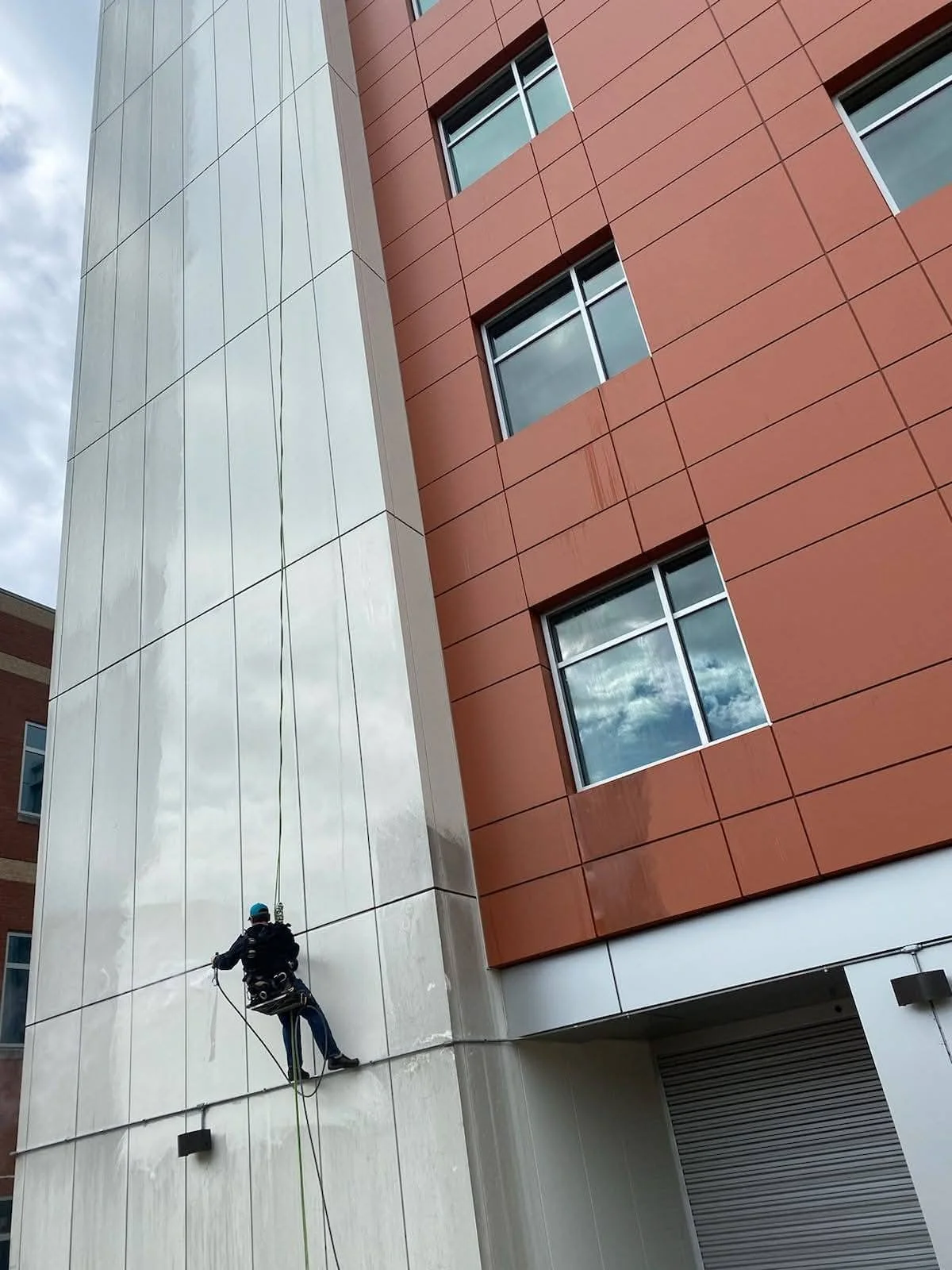 A worker wearing safety gear and a harness is cleaning or maintaining the exterior of a tall building, which has a combination of white and reddish-brown panels and several windows reflecting the cloudy sky.