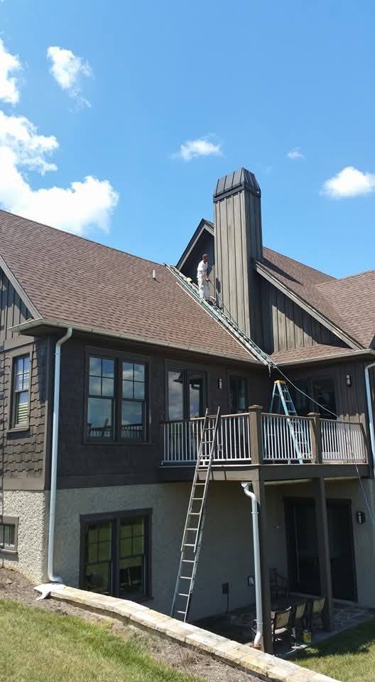 Person working on roof with ladder in front of house with chimney and balcony, under blue sky with clouds.
