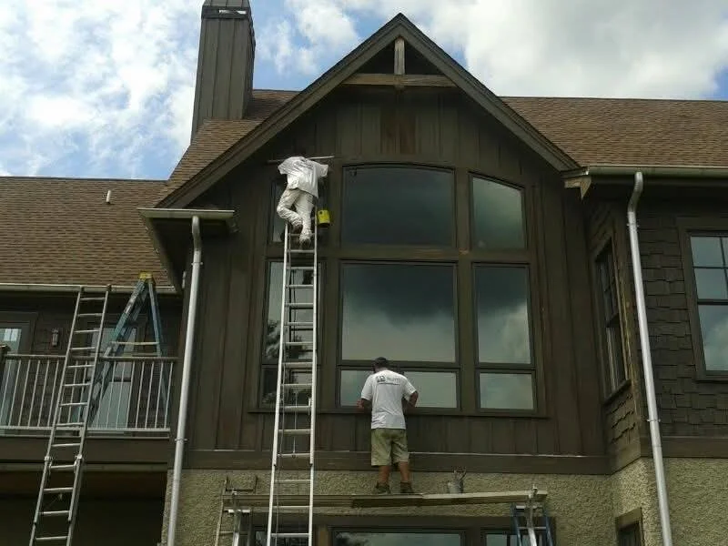 Two workers are painting or staining the exterior of a house, standing on ladders near large windows with a cloudy sky in the background.