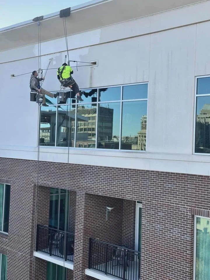 Workers cleaning windows on the exterior of a multi-story building using safety harnesses and scaffolding.