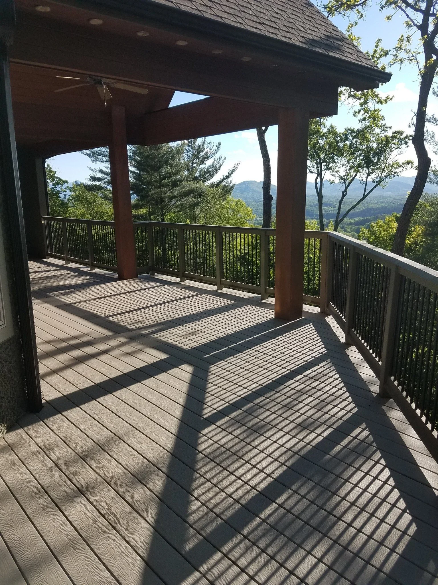 A spacious wooden balcony with railing, casting shadows from the sun, overlooking trees and distant mountains.