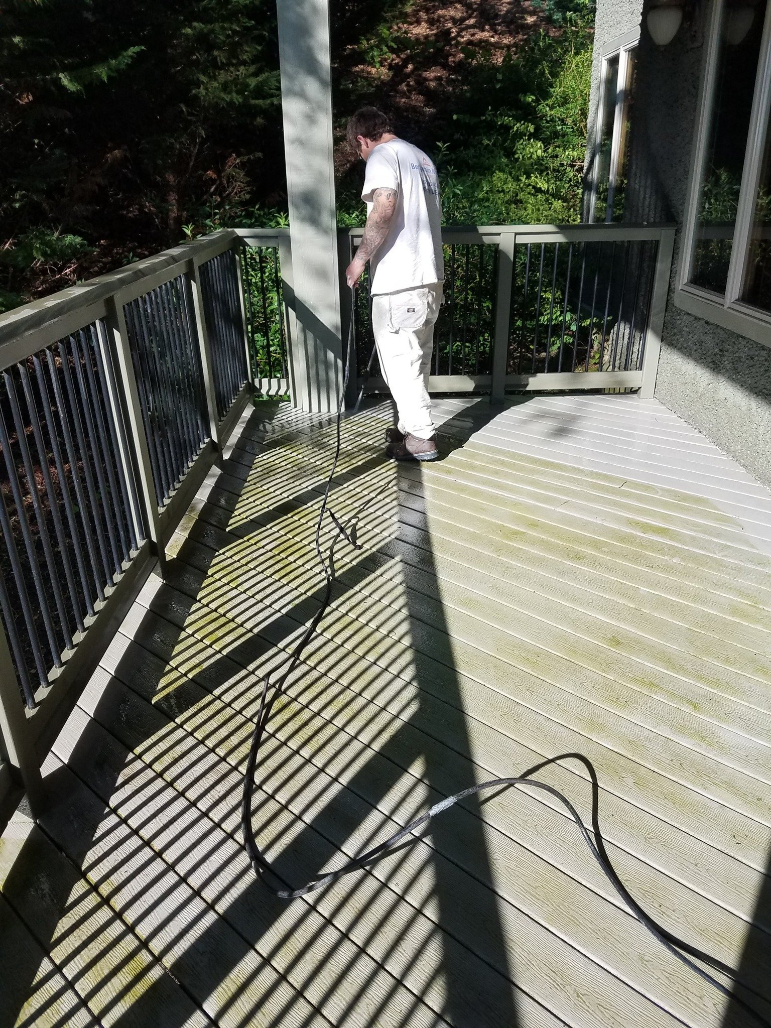A man power washing a wooden deck on an outdoor balcony, with shadows cast by the railing and the man.
