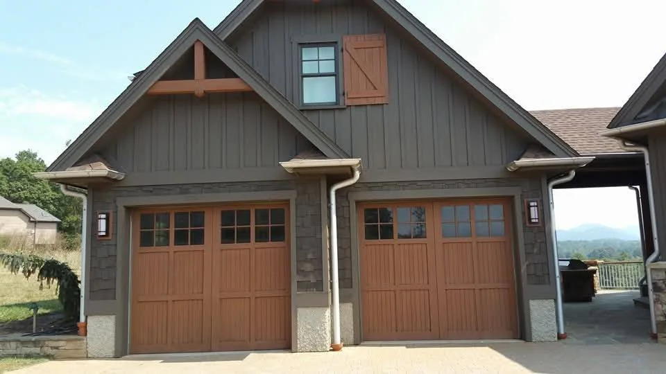 Front view of a modern two-car garage with wooden doors, gray siding, and small windows, attached to a house with outdoor lighting and a paved driveway.