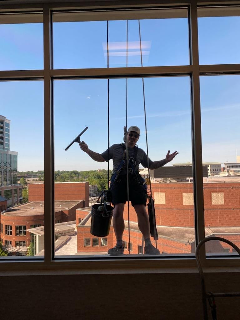 Window cleaner standing outside a tall building with a harness, holding a squeegee, and giving a peace sign.